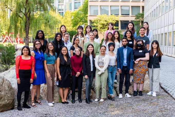 Nearly 30 students pose outdoors in between boulders and MIT Building 56 on a bright summer day.